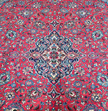 A close-up image of a traditional Persian Sarough rug featuring a geometric floral pattern in salmon pink, beige, and blue tones.