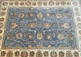 A traditional Afghan Chobi area rug with a dusty blue center field, decorated with beige, cream, and rust floral motifs, surrounded by a cream border.
