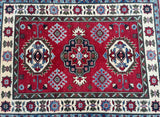 A rectangular, handmade, multicolored wool rug with a geometric Kazak pattern, featuring a combination of red, beige, blue, black, and green tones, and bordered with a decorative pattern.