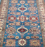 A close-up image of a hand-knotted Afghan Super Kazak rug with a geometric pattern in blue, beige, and red colors.