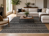A large rectangular contemporary Afghan area rug with a black and beige geometric tribal pattern is styled in a modern living room with a light beige sectional sofa, wooden coffee table, and potted plants.