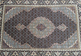 A finely woven Persian Tabriz rug with an intricate geometric pattern in beige, black, gold, green, and orange tones.
