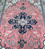 A vintage Persian Ardebil rug with a geometric pattern featuring a crossword-like design in a salmon pink, beige, black, blue, and brown color palette on a white background.