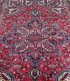 A traditional Persian Heriz area rug with a vibrant red field, featuring a large central medallion and intricate geometric and floral motifs in navy blue, beige, and white.