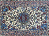 A traditional Persian Isfahan area rug signed with an intricate floral and medallion pattern, featuring a cream center, blue corners, and red and green accents, bordered by ornate pink and blue bands.