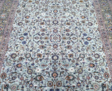 A close-up view of a signed Persian Kashan rug featuring a dense floral pattern with beige, brown, pistachio, and blue tones.