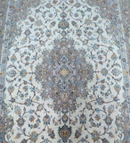 A traditional oriental Kashan area rug with an intricate floral and vine pattern, featuring a central medallion and border in muted tones of blue, gray, beige, and brown on a light cream background.