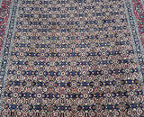 A close-up view of a signed Persian Mashad rug featuring an all-over Herati design with indigo blue, beige, red, orange, pink, and mustard tones on a dark background.