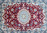 A hand-knotted Persian Nain rug with a central medallion floral design, featuring a colorful border in red, beige, blue, brown, and black, on a white background.