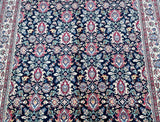 A close-up of an Persian Varamin rug with a dark navy blue field, featuring repeating floral and medallion motifs in red, pink, beige, and light blue, bordered by detailed red and cream patterns on the sides.