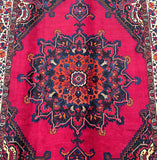 A deep maroon red Persian tribal rug with a central medallion pattern and floral accents, hand-knotted on a cotton base.