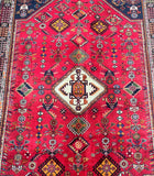 A close-up image of a hand-knotted Persian Qashqai Shiraz rug featuring a geometric multiple medallion design with red as the dominant color and accents in blue, beige, green, gold, black, and orange.