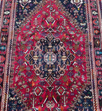 A close-up image of a Persian Qashqai Shiraz rug featuring a central medallion geometric design with animal motifs, predominantly in red, beige, blue, and black tones.