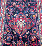 A close-up of a traditional patterned rug with a dark navy blue field, a large red central medallion, and intricate geometric and floral motifs in red, pink, beige, and blue.