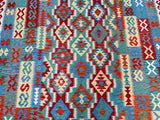 A decorative full-room size hand woven Afghan kilim rug with a geometric tribal design featuring a variety of colors including red, blue, green, beige, and brown.