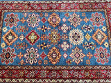 A hand-knotted Afghan Royal Kazak rug featuring a geometric design with blue, beige, orange, green, and maroon tones on a red and blue background.