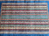 A vibrant, multicolored hand-knotted Afghan Super Kazak rug laid out on a blue background, showcasing its intricate stripe design and traditional patterns.
