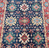 A close-up image of a hand-knotted Afghan Serapi Heriz rug featuring a geometric pattern with red, blue, green, and beige colors on a dark background.
