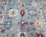 A multicolored handmade Afghan Ushak rug with a floral and geometric pattern, featuring a mix of light grey, beige, blue, soft orange, maroon, and purple tones.