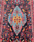 A vibrant, multicolored Persian Hamedan rug with a geometric and floral pattern, featuring a deep indigo blue/black, red, gold, light blue, and brown tone.