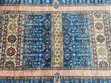 A handmade Afghan rug with a floral design in blue, beige, green, brown, caramel, and orange tones, featuring intricate patterns and a pile texture.